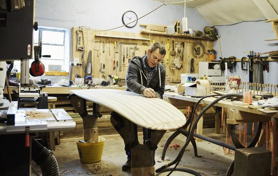 Man standing in a workshop sanding and shaping a wooden surfboard.