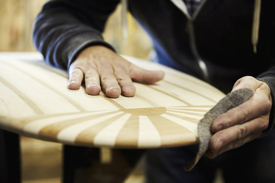 Man in a workshop sanding the edge of a wooden surfboard with a surface pattern of inlaid wood.