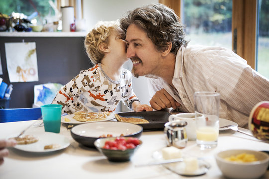 Boy whispering to fathers ear at kitchen table