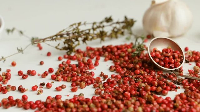 Pink peppercorns on white table with garlic bulbs, thyme, rosemary and spoon. Slow pan. 
