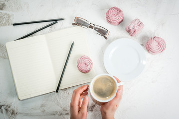 Home spring vacation, relax, work. White stone concrete desk with notepads, coffee and pink berry pastries (marshmallows). The girl holds a cup in his hands top view copy space