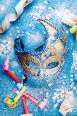 Carnivalesque composition. Top view of a carnival mask, whistles, corianders, and sfrappoles or chiacchere,italian fried carnival cookies with icing sugar on a bluish shabby chic board.