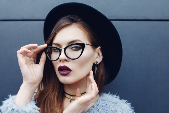 Young beautiful fashionable woman with long hair, trendy makeup. Model looking at camera, wearing stylish eyeglasses, fake fur coat, hat, accessories. Female fashion, beauty concept. Outdoor. Close up