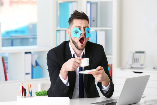 Young business man with fake eyes painted on paper stickers yawning at workplace in office