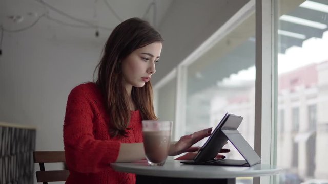 Young woman sits at a table at a restaurant using a tablet with access to the Internet, checking the mail and drinking coffee.