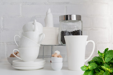 Tea drink concept. Piece of cafe, restaurant interior with teapot, tea cups, sugar cubes, tea leaves, paper napkins, saucers on white board against white brick wall with a bunch of green fresh mint.