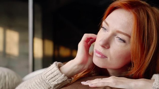 Portrait of young ginger woman lying on the bed and dreamily looking through a window