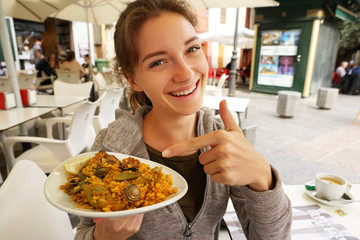 Paella - Spanish local food. Smiling woman points a finger at a plate of paella, at outdoor restaurant. Travel in Spain.