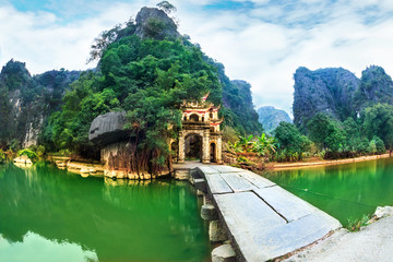 Fototapeta premium Outdoor park landscape with stone bridge and lake. Gate entrance to old Bich Dong pagoda complex. Ninh Binh, Vietnam - travel destination.