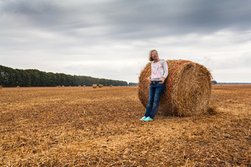 Naklejka na ściany i meble A young woman stands on the field, leaning on a sheaf of hay against the background of dark dramatic sky with clouds
