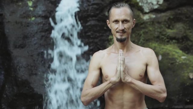  Young man practicing yoga fitness exercises outdoors against the backdrop of a beautiful waterfall. Meditation and relaxation. 
