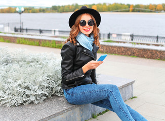 Fototapeta premium Fashion young smiling woman using smartphone wearing black rock jacket, hat in city park