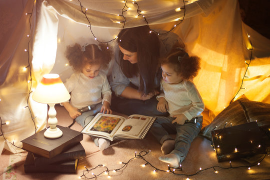 Reading and family games in children's tent. Mother and two twins daughters with books and flashlight before going to bed.