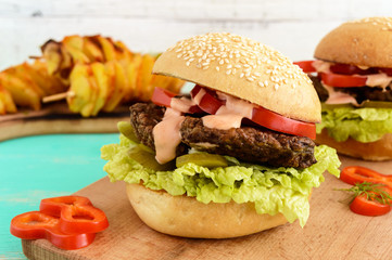 Burgers with liver cutlet, tomatoes, pickles, lettuce, spicy sauce and a soft bun with sesame seeds on a cutting board and potato slices on skewers. Close up