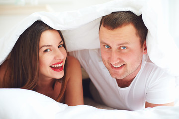 happy couple playing under duvet cover at bedroom