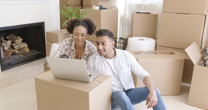 Happy mixed race couple sitting on the floor among unpacked carton boxes and using laptop computer to plan the future in their new apartment they just moved in.