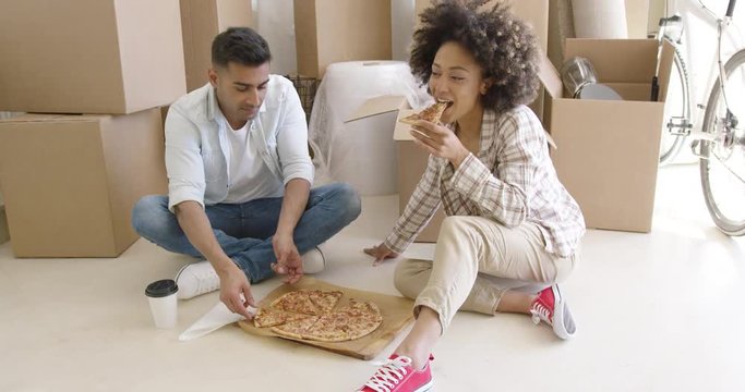 Young African American couple tucking into a pizza as they sit relaxing on the floor in front of a pile of packing boxes