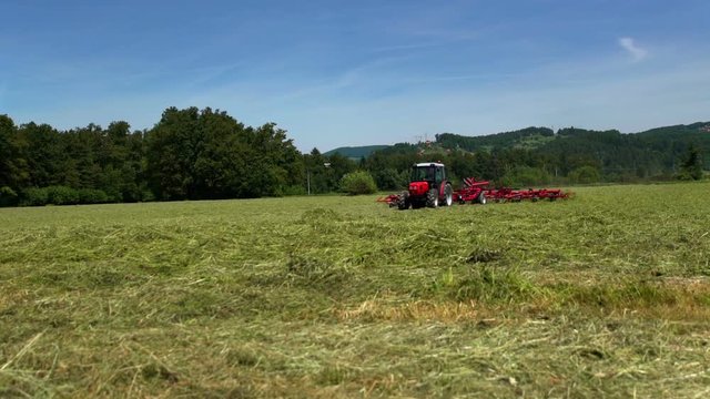 A big tractor is preparing hay on a grass field in summer time. There is a beautiful green forest in the background and the sun is shining.
