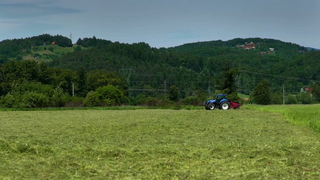 A blue tractor is making a turn on a large field and he is driving into the opposite direction. Farmers are preparing hay in summer time.
