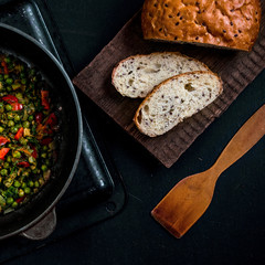 Vegetable stew with green peas, red pepper, onion and zucchini and spices. In a frying pan on a black background. On a wooden platform grain bread with flax seeds. Size 1 to 1.