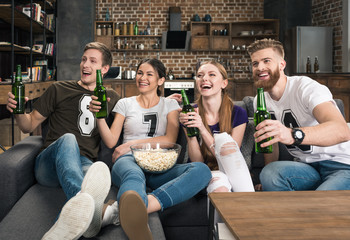 Cheerful young friends holding beer bottles and looking away at home