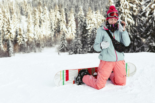 Woman in red hat, ski glasses and warm suit poses with snowboard on the hill