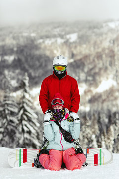 Woman on snowboard sits on the ground while man in red ski suit hugs her from behind on mountain hill