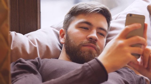 Smiling Bearded Man lying on sofa near the window in cafe and using smartphone