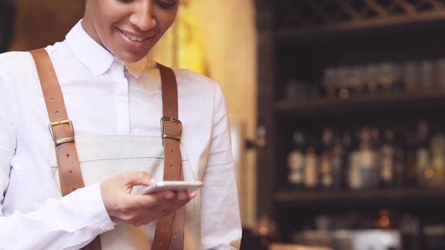 Dark-skinned smiling waitress looking back and using smartphone