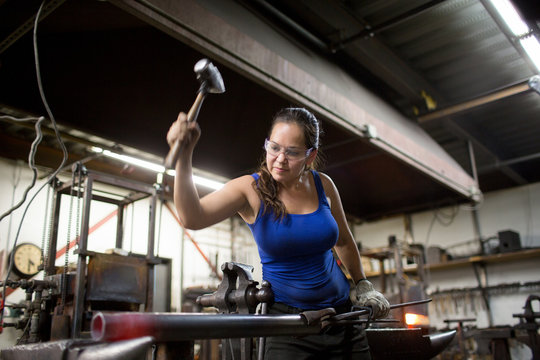 Female metalsmith hammering red hot metal rod on workshop anvil