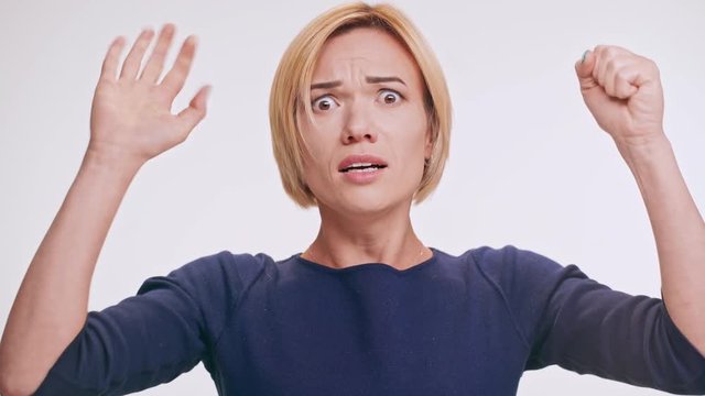 Scared middle-aged Caucasian blonde female in blue sweatshirt playing pantomime knocking on imaginary wall on white background