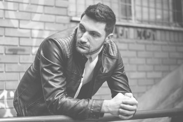 Handsome man with the beard and hairstyle posing in his clothes wearing a leather jacket and white t-shirt. in urban environment leaning against brick wall