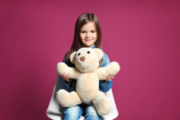Cute little girl with teddy bear sitting on chair against purple background