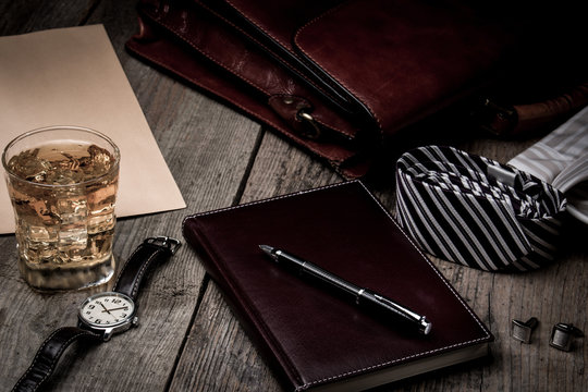 business accessories - notebook, pen, rich portfolio, watch, tie, glass of whiskey with ice on wooden background,
