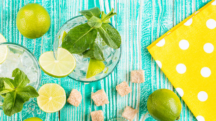mojito in glasses with ice cubes,  decorated by mint leaf, lime fruits, yellow napkin at white polka dots on turquoise colored wooden table, top view - 137283104