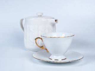 Picture of the white infuser teapot near tea cup decorated witn white and golden colour. Infuser teapot and tea cup standing on a saucer on white background.