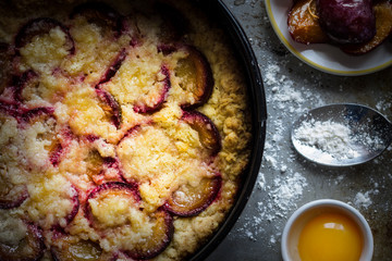 Plum Cake with Raw Cooking Ingredients on Wooden Kitchen Table. Dark Food Photography.