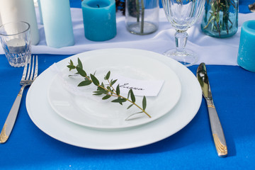Plates with greenery on the wedding banquet table with blue tablecloth and cutlery, glasses, candles