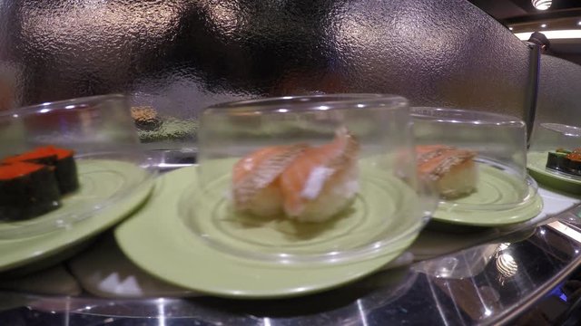 4K, Asian Woman Taking Sushi On Conveyor Belt In Japan Restaurant. Sushi is a food preparation with raw uncooked seafood originating in Japan. Sashimi is often served with wasabi and soy sauce-Dan