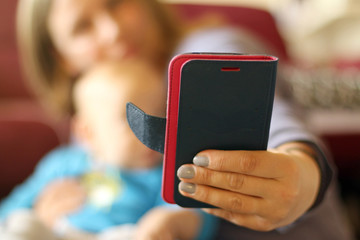 Mobile phone in mom's left hand while is making selfie with baby in the arms