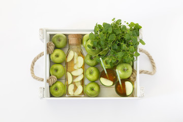 Top view of a ripe green apples in a white wooden box with mint and lemon balm. Apple juice in a glass bottle and a glass with a straw on a light background.