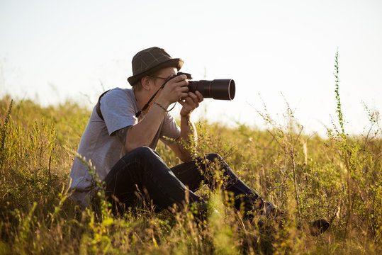Young male photographer in hat taking picture, sitting in field.