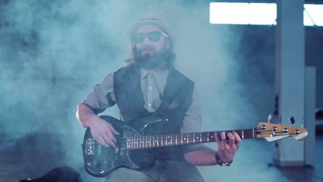 Middle shot of bearded man in sunglasses and hat wearing vest and shirt sitting on chair, playing black electric guitar and smiling in empty hall with studio lighting. Camera moving around