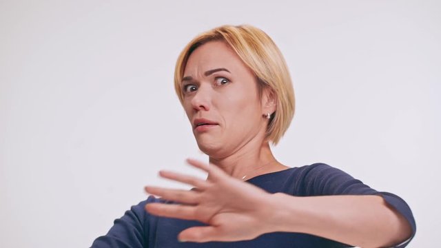Scared middle-aged Caucasian blonde female in blue sweatshirt playing pantomime pushing away on white background in slowmotion