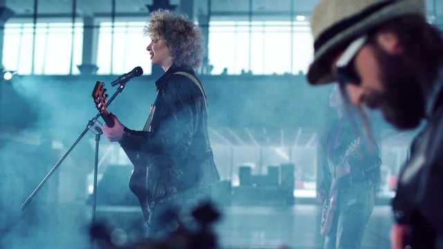 Rock band with curly young woman lead singer performing in empty hall, singing and dancing while shooting music video with smoke. Side view with male guitar player in foreground. Movement round shot