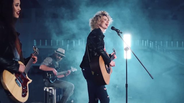 Smiling woman singing and playing the acoustic guitar while video production process. Camera moving around 4K