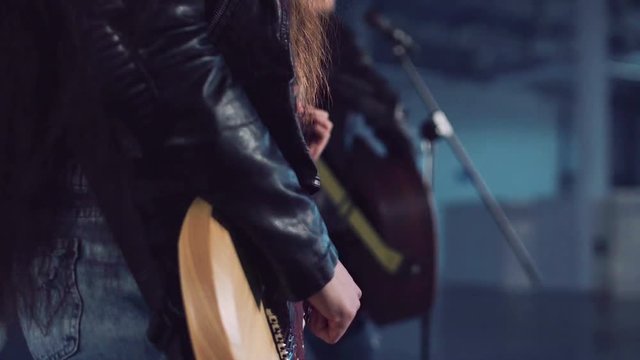 Cropped clip close shot of young incognito woman in leather jacket and ripped jeans playing electric guitar and dancing in empty hall. Camera moving around