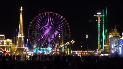 People are having fun in theme park with Ferris wheel at night. Ferris wheel shine with lots of colors and crown is walking around.