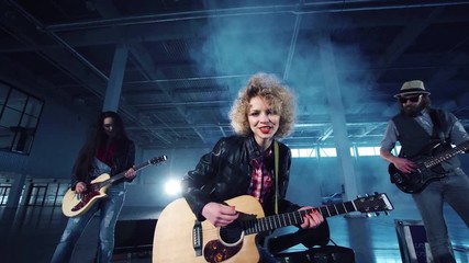 Low angle camera quickly zooming in shooting rock band playing guitars with curly woman in leather jacket singing and two men standing on sids. Indoors empty dark hall - Powered by Adobe