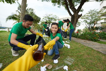 Joyful Asian woman sitting on haunches and picking up glass bottles in blooming park